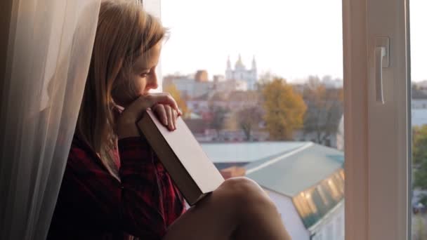 Όνειρα νεαρή γυναίκα με το βιβλίο που κάθεται σε ένα windowsill στο σπίτι — Αρχείο Βίντεο