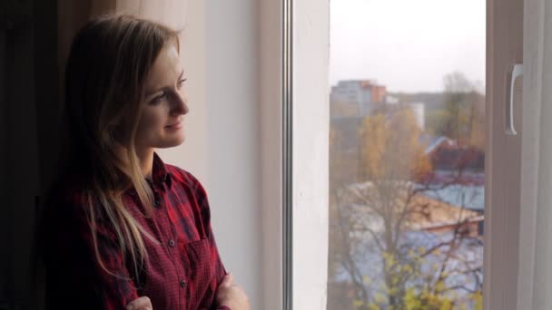 Vacker ung kvinna med bok tittar ut genom fönstret och ler — Stockvideo