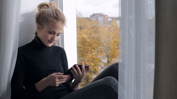 Όμορφη γυναίκα διαβάζοντας ένα βιβλίο στο e-reader που κάθεται σε ένα windowsill — Αρχείο Βίντεο