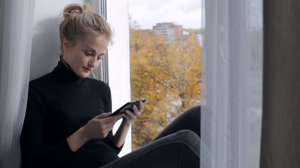 Женщина сидит на подоконнике и читает книгу в электронном читателе — стоковое видео