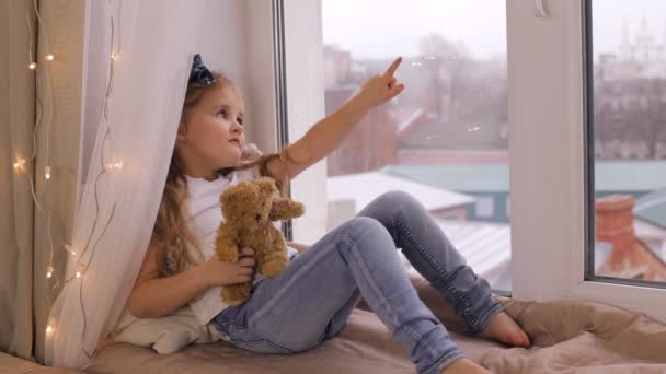 Linda chica jugando con su oso de peluche y sentado en el alféizar de la ventana — Vídeos de Stock