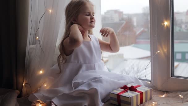 Ładna dziewczyna siedzi na parapecie ozdobione Boże Narodzenie girlanda i rozpakowaniu jej obecny — Wideo stockowe