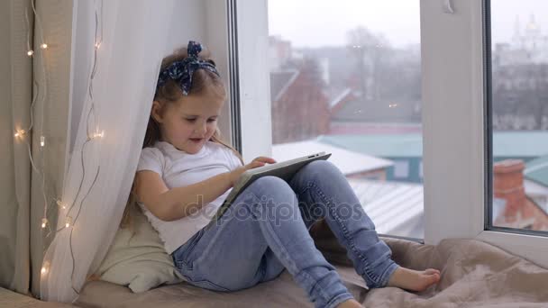 Liten flicka sitter på fönsterbrädan och hålla tabletten — Stockvideo