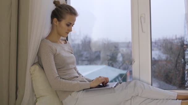 Hermosa mujer escribiendo en su computadora portátil y sentado en el alféizar de la ventana — Vídeos de Stock