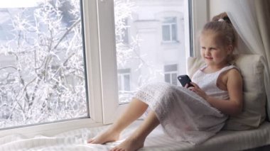 Genç kız onun smartphone üzerinde bir pencere pervazına müzik dinler. Güneşli kış sabahı