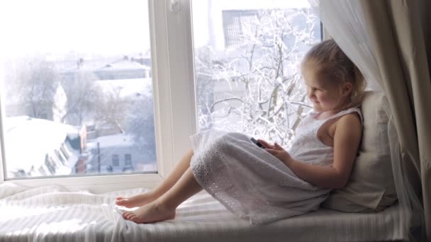 Lustiges junges Mädchen mit Smartphone auf der Fensterbank. sonniger Wintermorgen — Stockvideo