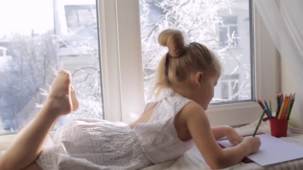 Malá holčička kreslí obrázky ležící na okenním parapetu. Slunečné zimní ráno — Stock video