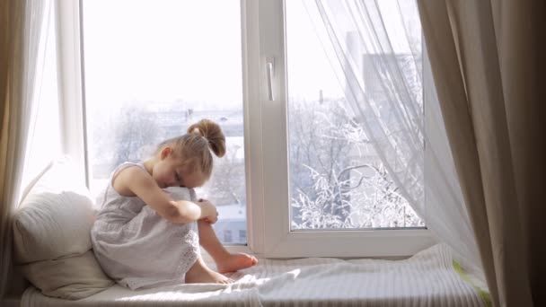 Niña triste sentada en el alféizar de la ventana. Invierno fuera de la ventana — Vídeo de stock