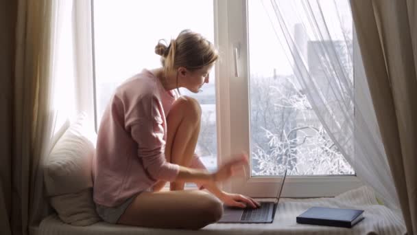 Frau arbeitet an ihrem Laptop und sitzt auf Fensterbank am Fenster — Stockvideo