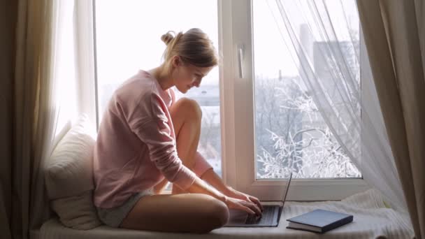 Fiatal nő ül, ablakpárkányon laptop. Majd ő keres az ablakon. — Stock videók