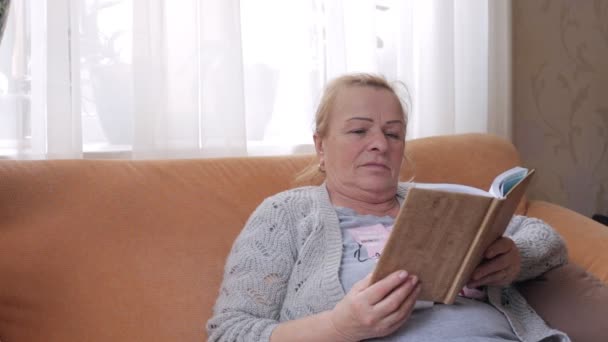 Старшая женщина сидит на диване и читает книгу — стоковое видео