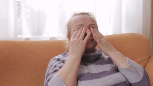 Elderly woman rubs her eyes while sitting on the sofa — Stock Video