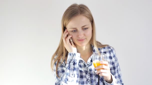 Молодая женщина пьет стакан апельсинового сока разговаривая на смартфоне — стоковое видео
