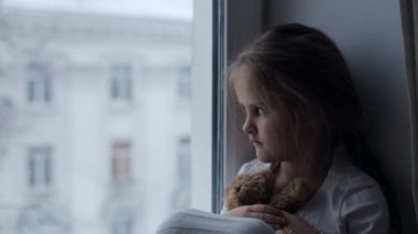 Güzel üzgün küçük kız ayısıyla pencere eşiğinde oturuyor.