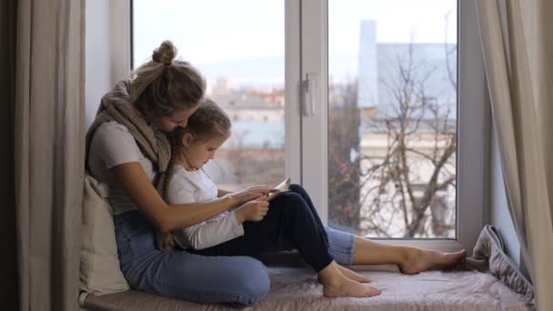 Mom with a small daughter are sitting on the windowsill and reading a book — Stock Video