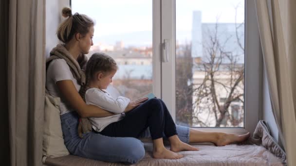 Mother and daughter are sitting on the windowsill and sadly looking out the window — Stock Video