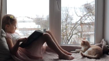 Küçük kız bir kediyle pencere kenarında oturur ve bir kitap okur.