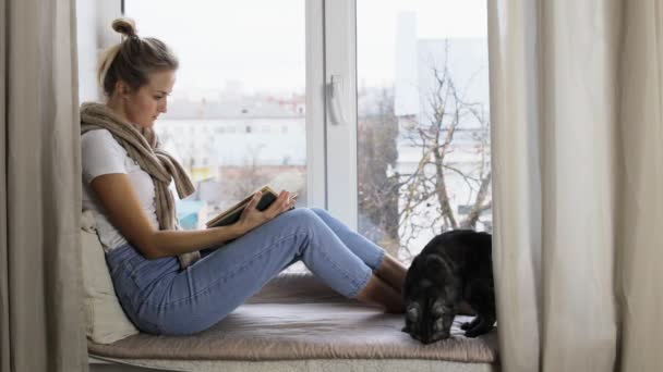 En flicka läser entusiastiskt en bok när hon sitter på en fönsterbräda — Stockvideo