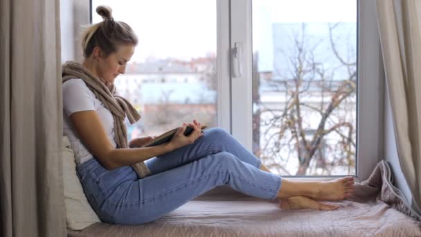 Een jong meisje leest een boek terwijl ze op een vensterbank zit — Stockvideo