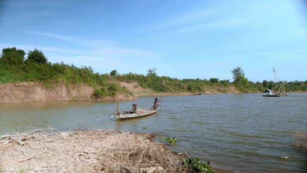 Fischer rudert mit einem Einbaum auf dem See; Hausboot mit chinesischem Fischernetz als Hintergrund (Blick vom Flussufer) — Stockvideo