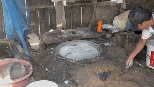 Dentro de un taller de fideos de arroz con agua hirviendo sobre fogones en el suelo. Hombre añadiendo virutas de madera para mantener el fuego — Vídeos de Stock