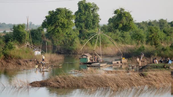 Pescador de la red de fundición de pequeñas plataformas de madera en un lago, mientras que la red de pesca china se levanta fuera del agua en el fondo — Vídeos de Stock