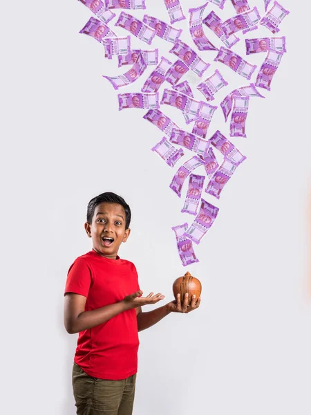 10 year old indian boy excited to see newly launched 2000 rupees indian currency notes falling in clay money box, asian boy holding piggy bank and money falling into it, indian currency flying — Stock Photo, Image