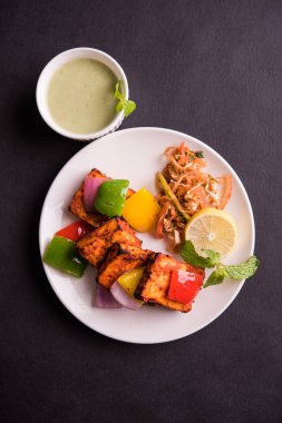 Paneer Tikka veya acı biber kebabı - Tandır Hint peyniri şişleri, beyaz tabakta servis edilir renkli kapsiyum, soğan ve yeşil sos ile