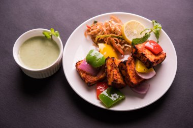 Paneer Tikka veya acı biber kebabı - Tandır Hint peyniri şişleri, beyaz tabakta servis edilir renkli kapsiyum, soğan ve yeşil sos ile
