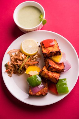 Paneer Tikka veya acı biber kebabı - Tandır Hint peyniri şişleri, beyaz tabakta servis edilir renkli kapsiyum, soğan ve yeşil sos ile