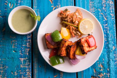 Paneer Tikka veya acı biber kebabı - Tandır Hint peyniri şişleri, beyaz tabakta servis edilir renkli kapsiyum, soğan ve yeşil sos ile