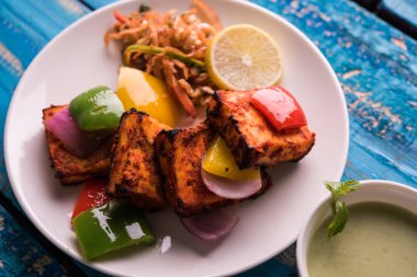 Paneer Tikka veya acı biber kebabı - Tandır Hint peyniri şişleri, beyaz tabakta servis edilir renkli kapsiyum, soğan ve yeşil sos ile