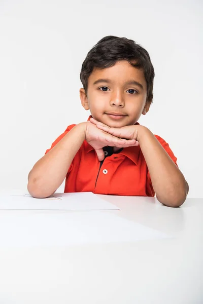 Happy indian gypsy child boy sitting at school table, isolated on white background, indian kid sitting at table with chin over both hands — Stock Photo, Image