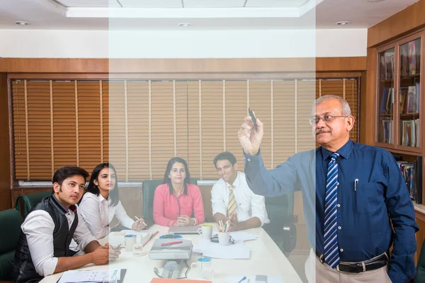 indian senior businessman writing over transparent glass with marker pen giving presentation in meeting room, while other young business people in the background