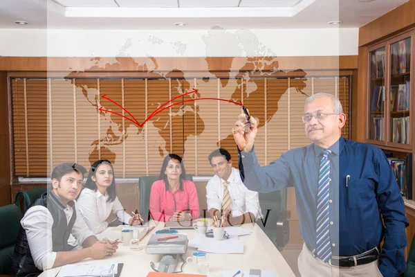 Empresario sénior indio escribiendo sobre vidrio transparente con rotulador dando presentación en la sala de reuniones, mientras que otros jóvenes empresarios en el fondo — Foto de Stock