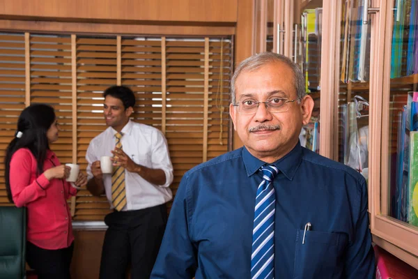 Indien beau homme d'affaires senior debout avec les bras dans la poche tandis que 2 jeunes hommes d'affaires prendre un café et discuter en arrière-plan — Photo