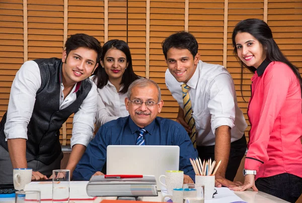 Satisfied with new plan. Group of four to five Indian cheerful young people and one experienced businessman looking at laptop with smile while leaning to the table in office Stock Image