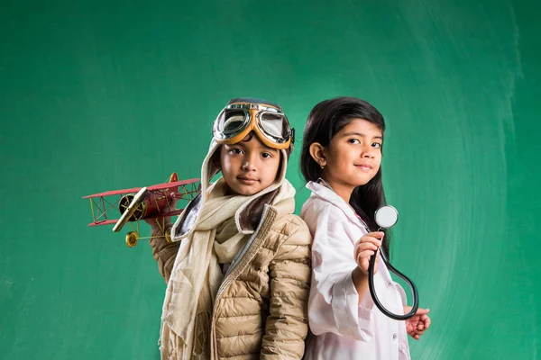 Kids and education concept - Small indian boy and girl posing in front of Green chalk board in pilot fancy dress and doctor costume with stethoscope, wanna pilot or doctor Royalty Free Stock Photos