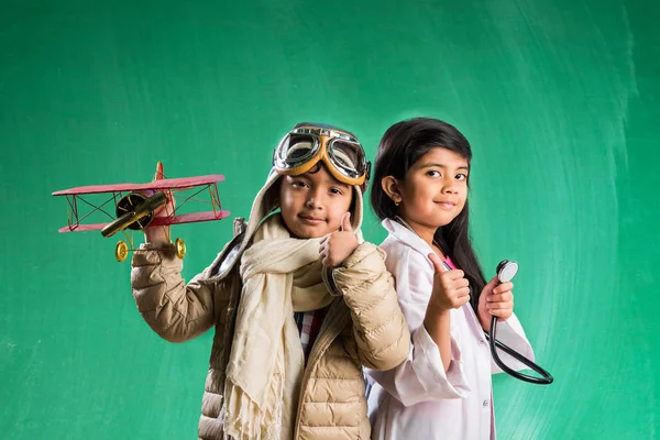 Kids and education concept - Small indian boy and girl posing in front of Green chalk board in pilot fancy dress and doctor costume with stethoscope, wanna pilot or doctor Royalty Free Stock Images