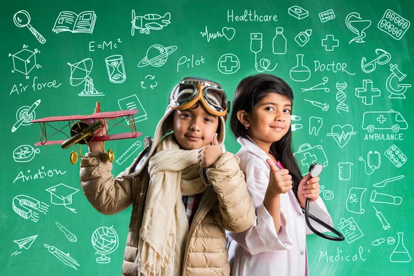 Kids and education concept - Small indian boy and girl posing in front of Green chalk board with doodles in pilot fancy dress and doctor costume with stethoscope, wanna be pilot or doctor Stock Picture