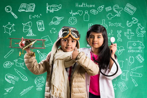 Kids and education concept - Small indian boy and girl posing in front of Green chalk board with doodles in pilot fancy dress and doctor costume with stethoscope, wanna be pilot or doctor Stock Picture