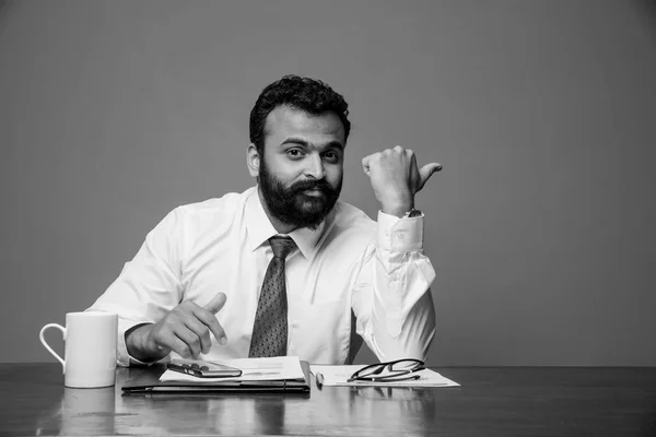 Homem de negócios indiano barbudo sentado à mesa e apresentando algo com a mão esquerda, apontando para a parede em branco com copyspace, asiático empresário apresentando algo — Fotografia de Stock