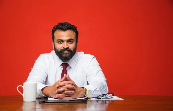 portrait of young Indian businessman in beard, sitting in relaxed position at at office table with pleasant look. Confident asian male business person