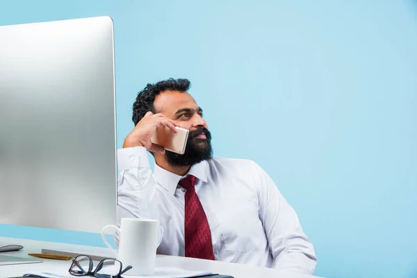 Young Indian businessman in beard busy on phone call while using laptop or computer in office, asian businessman talking using smartphone