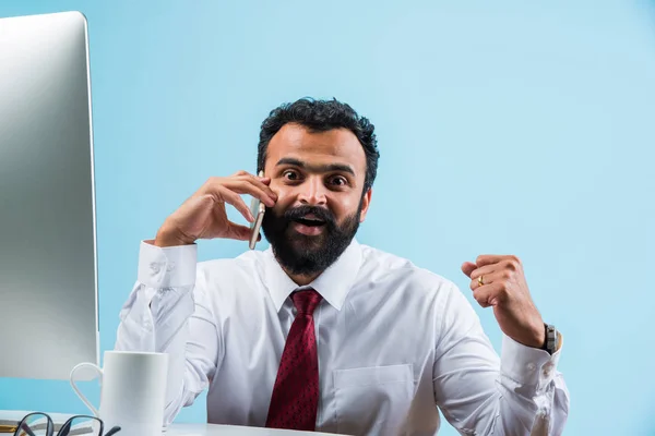 Unga indiska affärsmannen i skägg upptagen på telefonsamtal medan du använder bärbara eller stationära dator på kontoret, Asiatiska affärsman talar med smartphone — Stockfoto