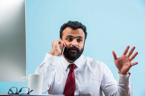 Jeune homme d'affaires indien dans la barbe occupé sur appel téléphonique tout en utilisant un ordinateur portable ou un ordinateur au bureau, homme d'affaires asiatique parlant à l'aide d'un smartphone — Photo