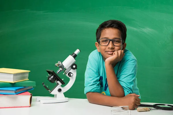 Kids and science concept - cute indian little boy studying science and using microscope with diagrams or doodles drawn over green chalkboard background