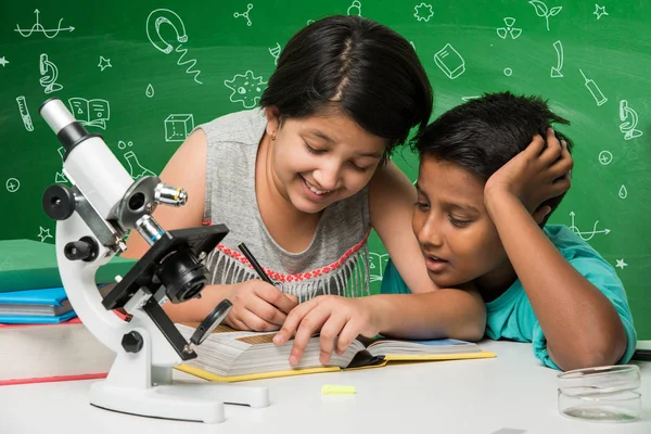 Kids and Science concept - Cute Indian little kids busy doing chemistry or science experiment with test tube and flask with safety eye glass over green chalkboard background with science doodles drawn