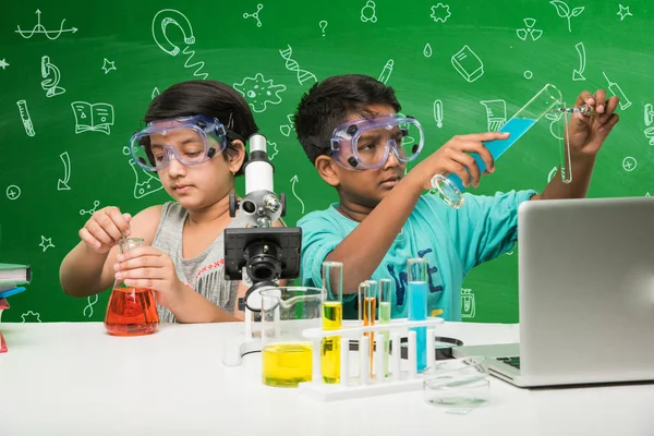2 little indian kids doing science experiment or chemistry experiment in classroom with safety eye glasses on standing over green chalkboard background with science doodles drawn over it
