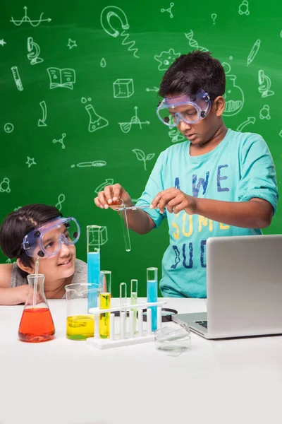 2 crianças indianas fazendo experimento de ciência ou experimento de química em sala de aula com óculos de segurança em pé sobre fundo de quadro-negro verde com doodles científicos desenhados sobre ele — Fotografia de Stock